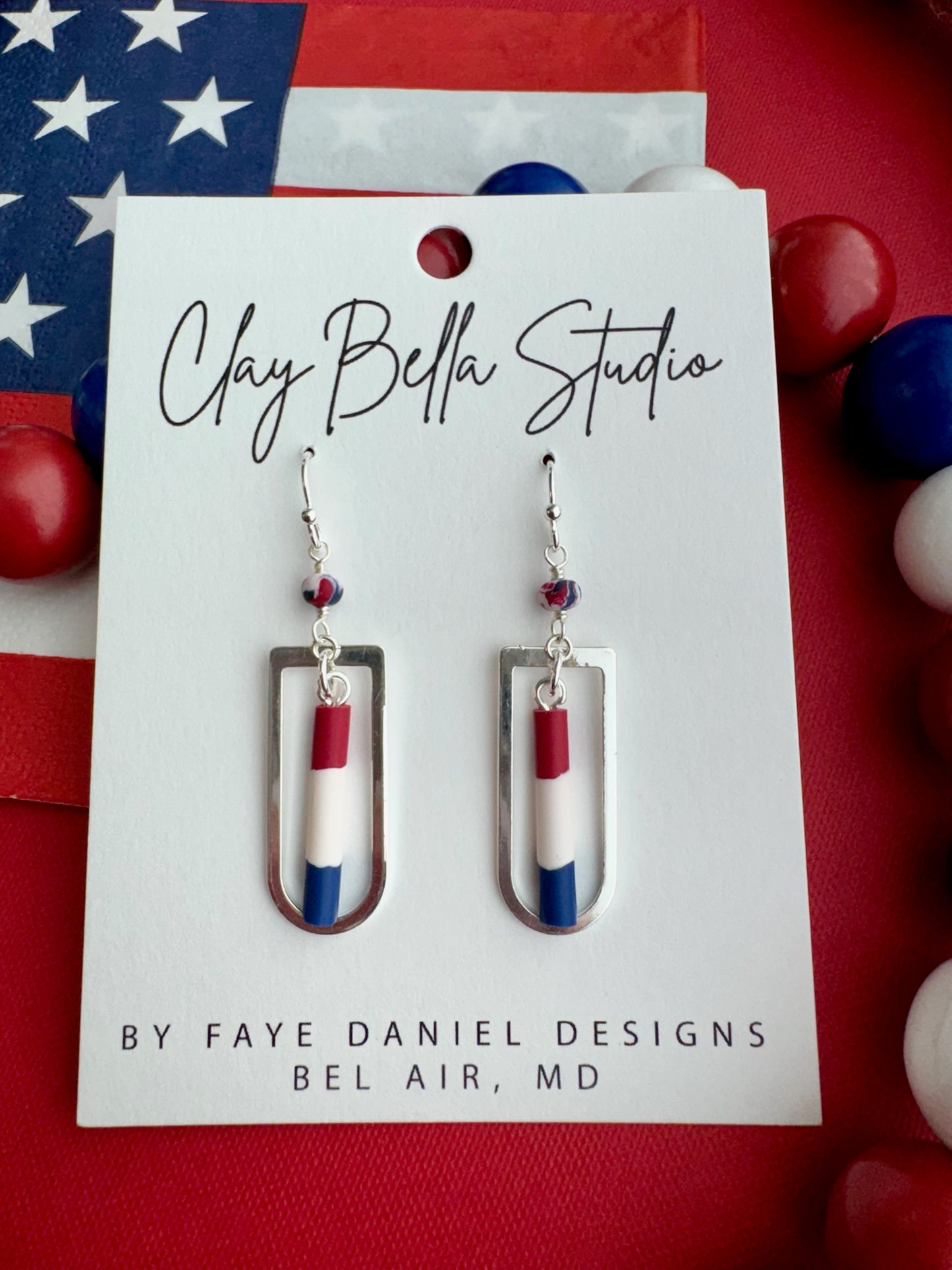 Handmade red, white, and blue polymer clay earrings suspended inside silver rectangular frames displayed on a Clay Bella Studio card against an American flag background.