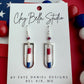 Handmade red, white, and blue polymer clay earrings suspended inside silver rectangular frames displayed on a Clay Bella Studio card against an American flag background.
