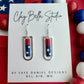 Handmade red, white, and blue polymer clay earrings  inside silver rectangular frame drops displayed on a Clay Bella Studio card against an American flag background.
