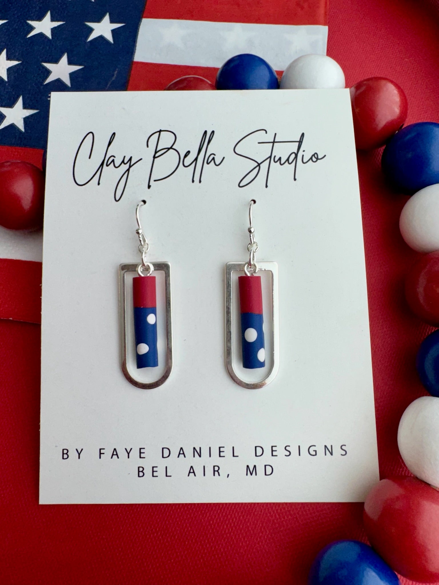 Handmade red, white, and blue polymer clay earrings  inside silver rectangular frame drops displayed on a Clay Bella Studio card against an American flag background.
