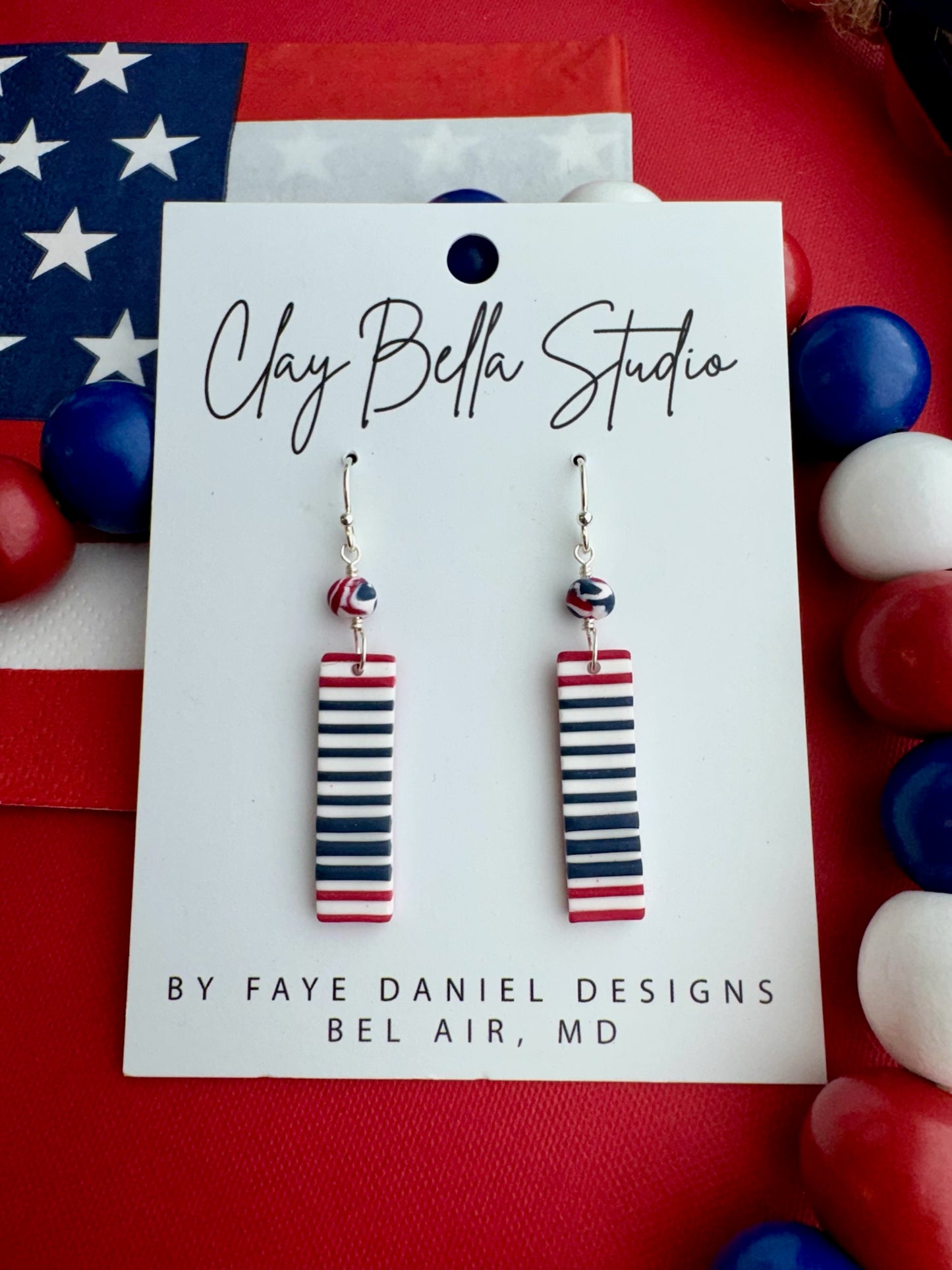 Red, white, and blue striped earrings on a Clay Bella Studio card with American flag in the background.