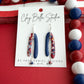 Red, white and blue asymmetrical polymer clay earrings with silver chain and bead accents on Claybella Studio display card.