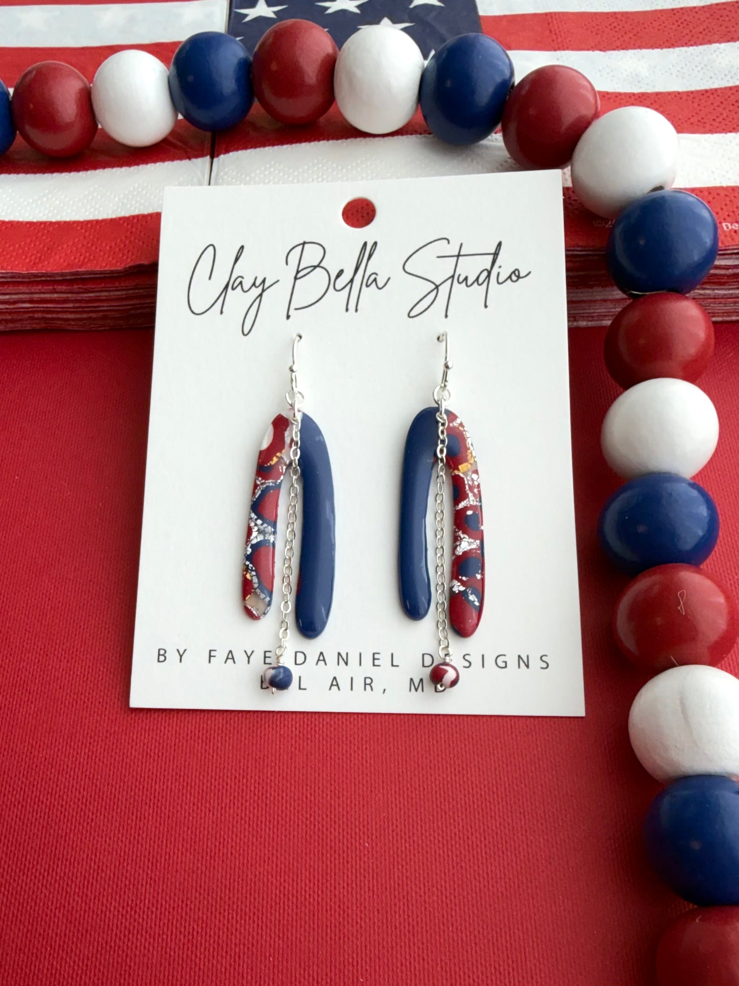 Red, white and blue asymmetrical polymer clay earrings with silver chain and bead accents on Claybella Studio display card.